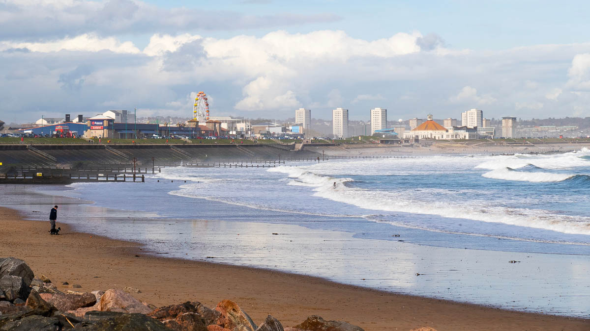 Aberdeen Beach looking south towards Codona's Amusement Park and the Beach Ballroom, with a dog walker on the sand and a surfer in the waves