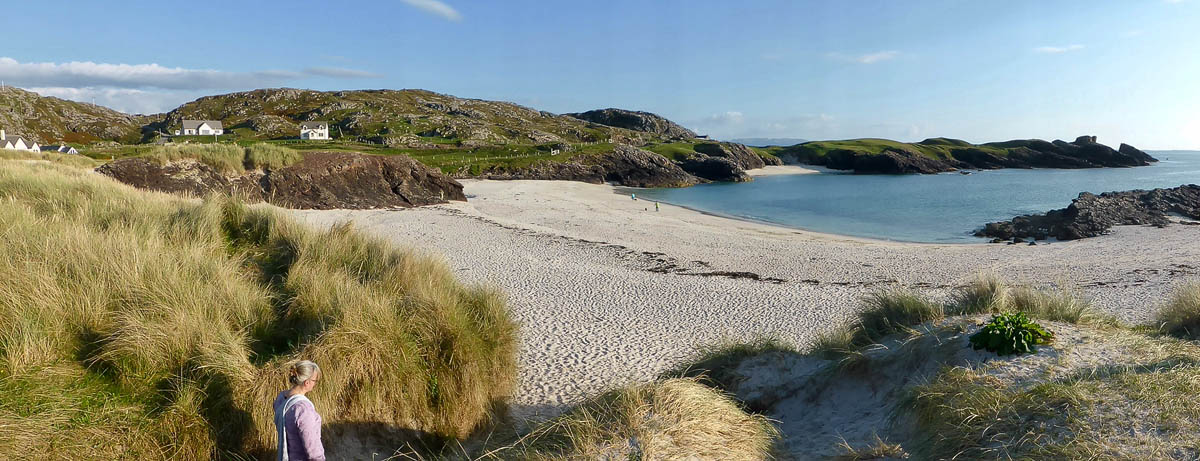 Panoramic view of Achmelvich Beach in Assynt with white sand, turquoise water, and a figure walking through marram grass dunes