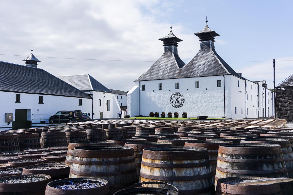 Ardbeg Distillery on Islay with its distinctive twin pagodas and Celtic knot logo, rows of whisky casks in the foreground