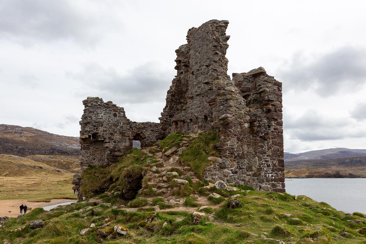 Ardvreck Castle ruins up close showing the crumbling walls and tower with visitors walking along the beach