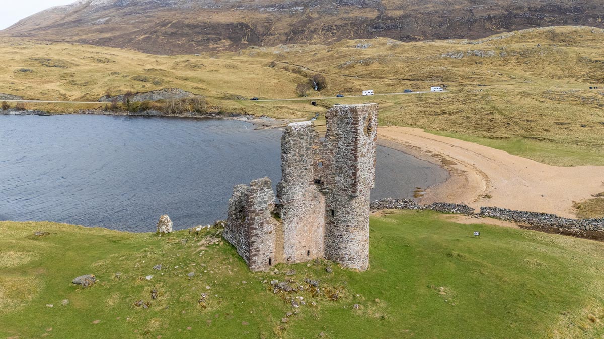 Aerial drone view of Ardvreck Castle showing the promontory, sandy beach, car park and the A837 road