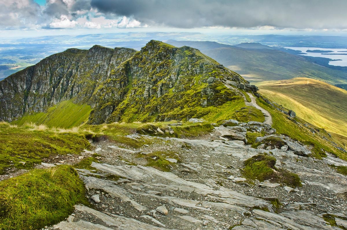 The rocky summit ridge path of Ben Lomond with dramatic cliff edges and Loch Lomond visible in the distance beyond rolling green hills