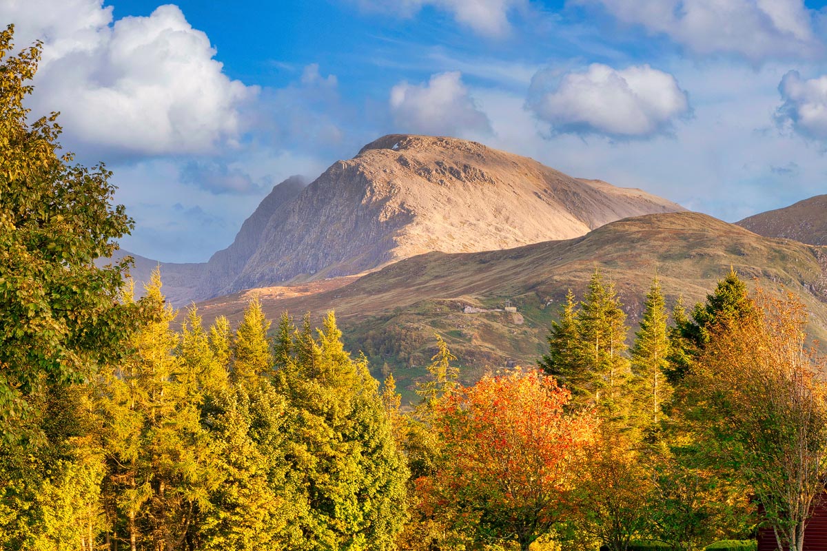 Ben Nevis, the highest mountain in the British Isles, seen from the approach with vibrant autumn-coloured trees in the foreground and blue sky above