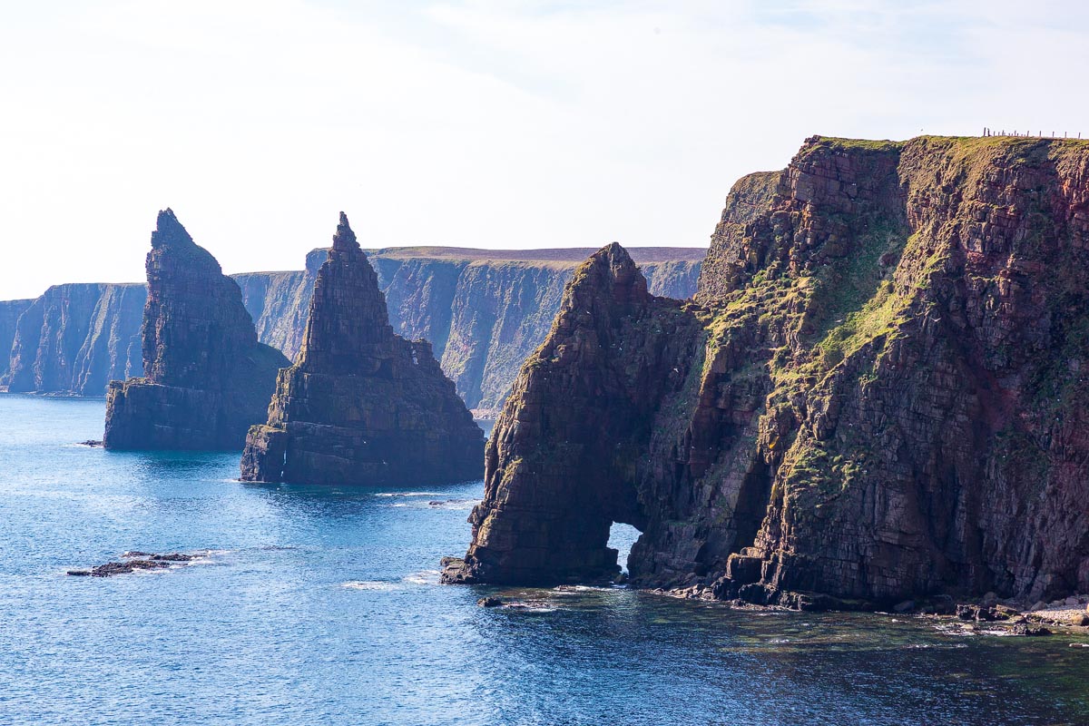 The Duncansby Stacks sea stacks rising dramatically from the blue sea with towering sandstone cliffs and a natural arch visible in the rock formations