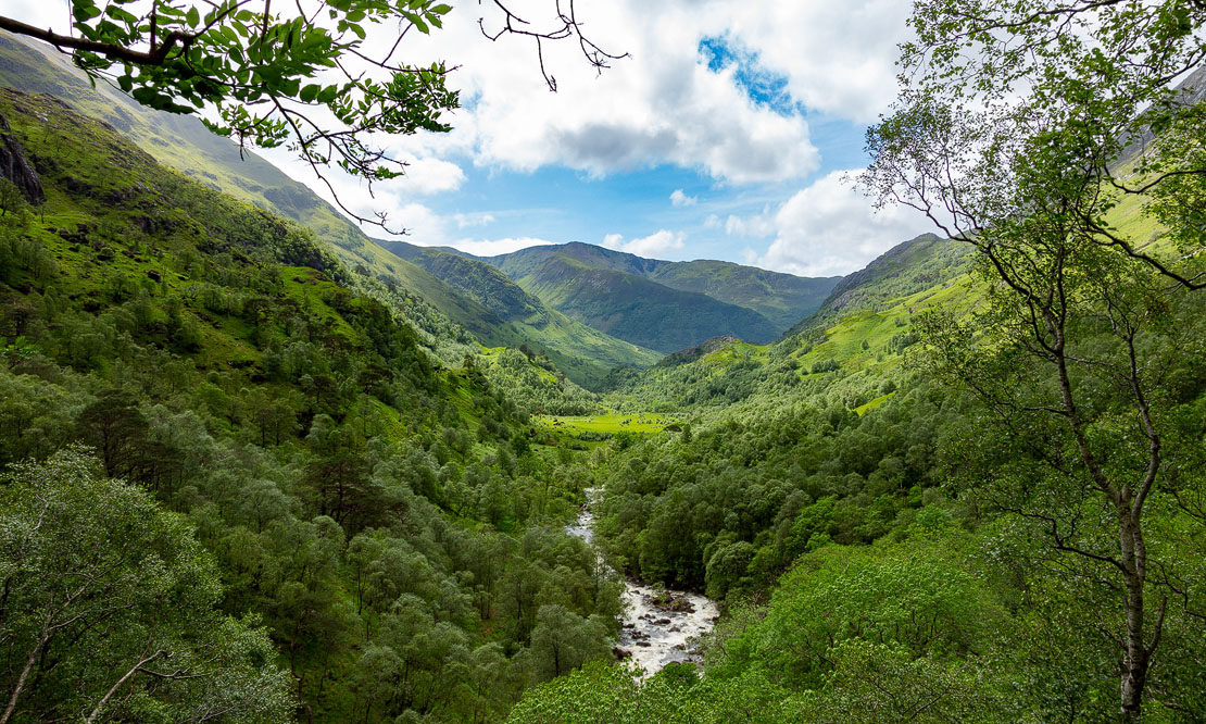 View through Glen Nevis gorge looking up the lush green valley with the Water of Nevis flowing through dense forest and mountains rising steeply on either side