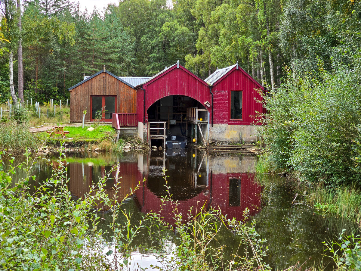 Red boathouse and timber-clad building reflected in the boat channel at Loch of Blairs