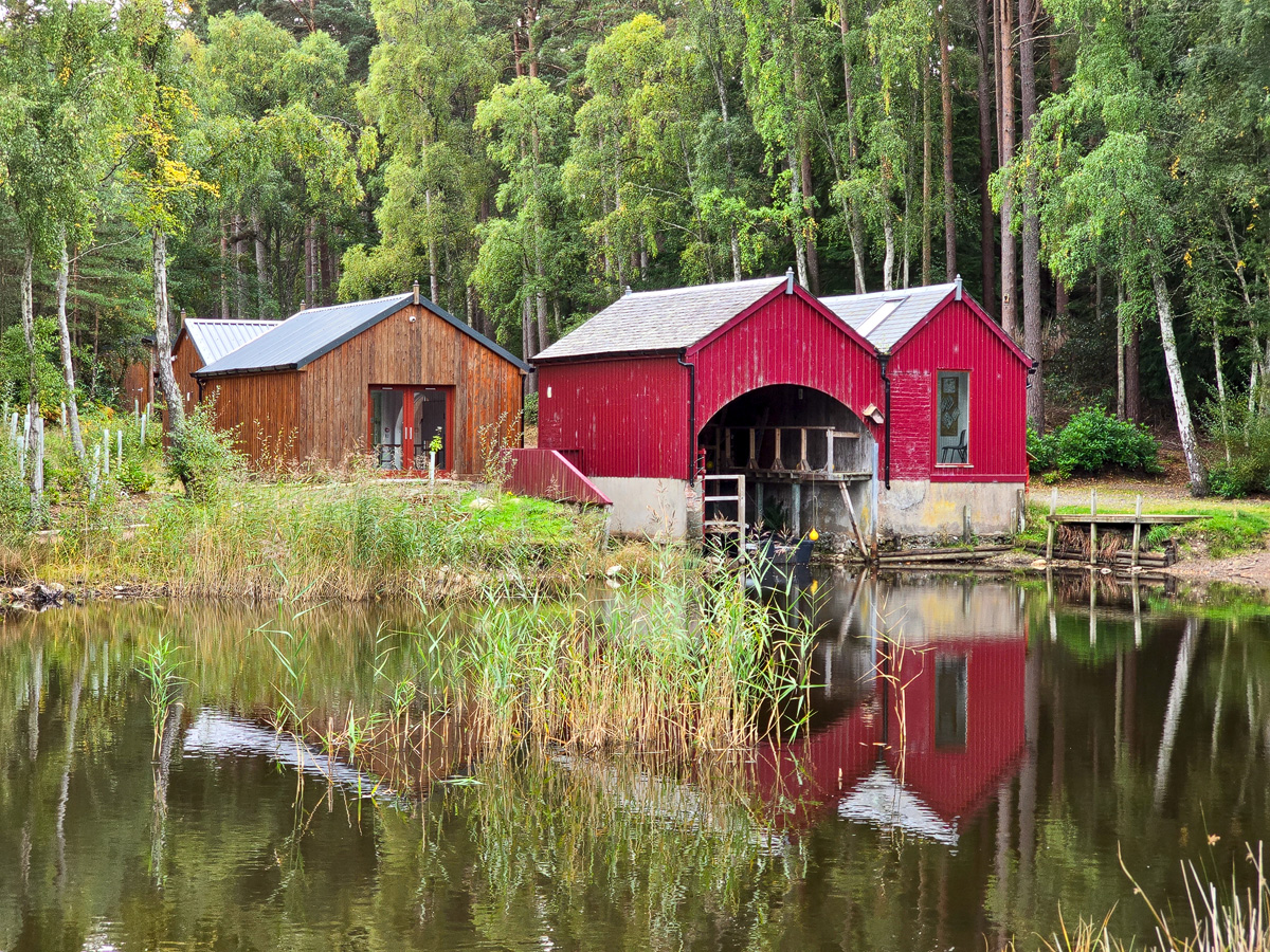 The red boathouse and bothy at Loch of Blairs reflected in still water, surrounded by birch and pine