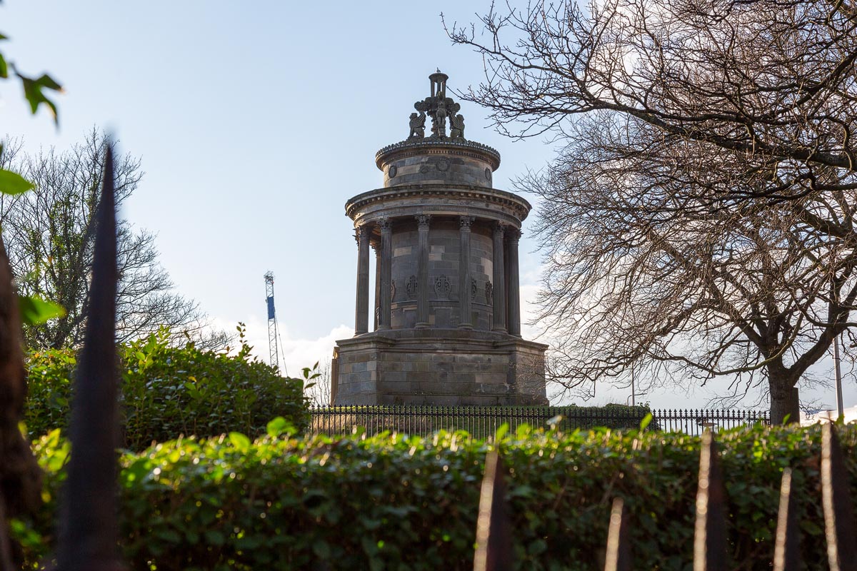 The Burns Monument in Edinburgh viewed through the garden hedgerow and iron railings, framed by bare winter trees