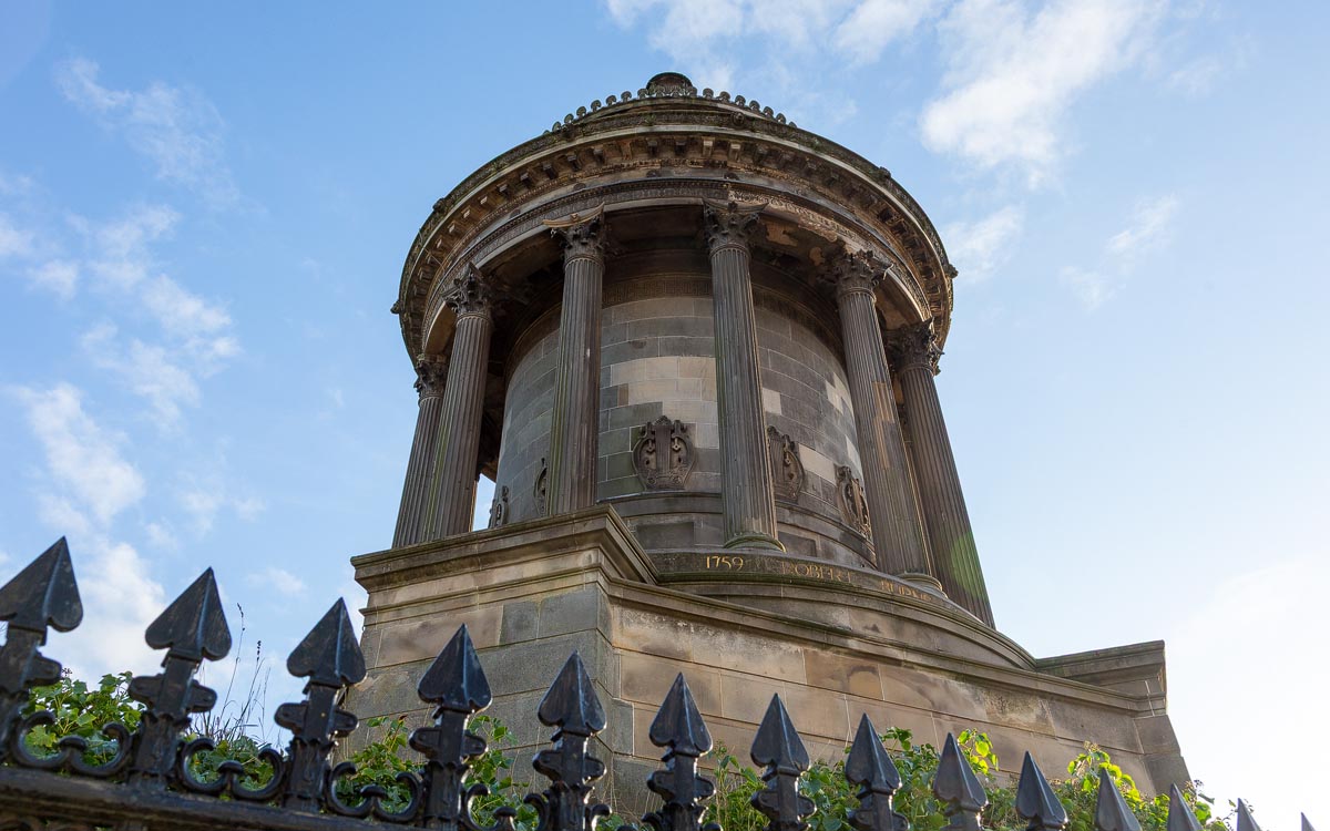 The Burns Monument viewed from below through ornate iron railings with fleur-de-lis finials, the gold inscription partially visible on the base