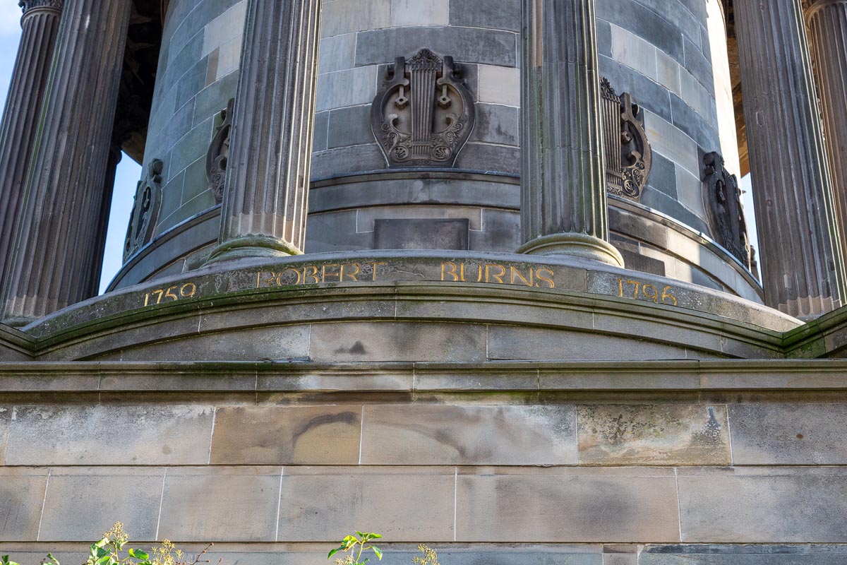 Close-up of the gilded inscription reading 1759 Robert Burns 1796 on the base of the Burns Monument, with carved lyre motifs visible above