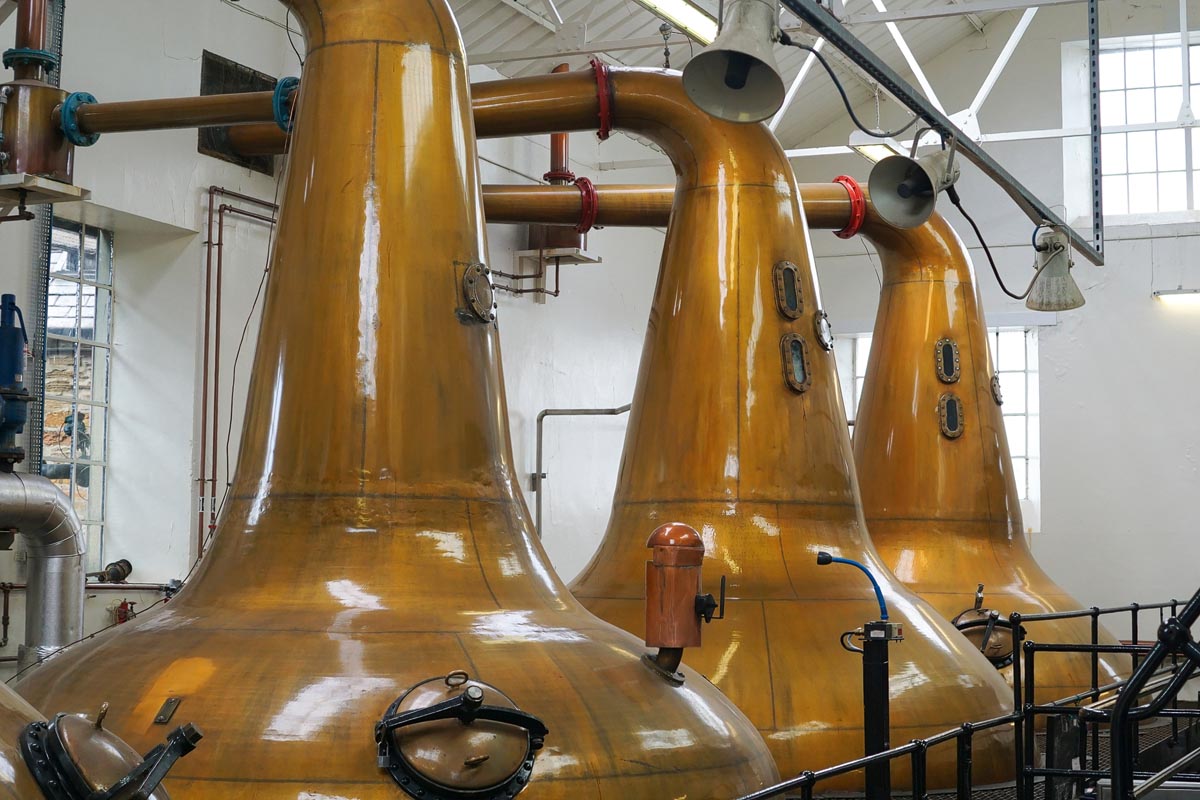Three large copper pot stills in a traditional Scottish distillery stillhouse with sight glasses and lyne arms visible