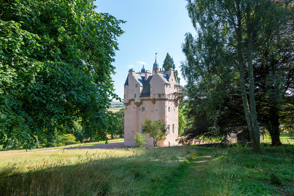 Craigievar Castle glimpsed through mature trees on the woodland walk with dappled sunlight and rolling countryside beyond