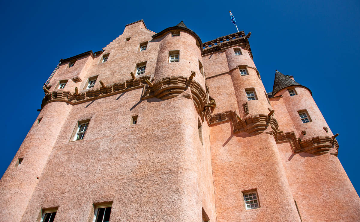 Looking up at Craigievar Castle from ground level showing corbelling turrets and pink harling texture with Saltire flag flying