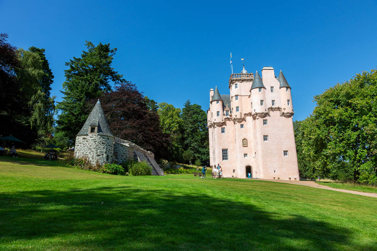Wide view of Craigievar Castle and the stone barmkin outbuilding with pointed roof showing the castle entrance and green lawns