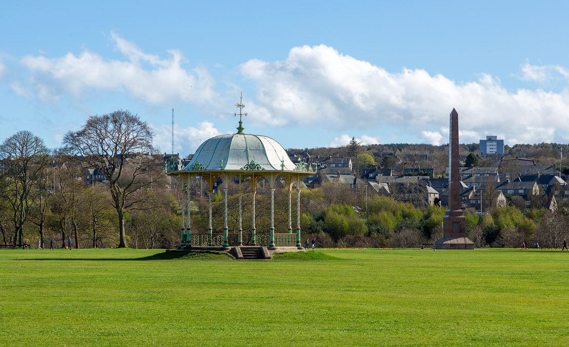 Distant view of the Duthie Park bandstand set on the broad central green with the McGrigor Obelisk and Aberdeen skyline behind