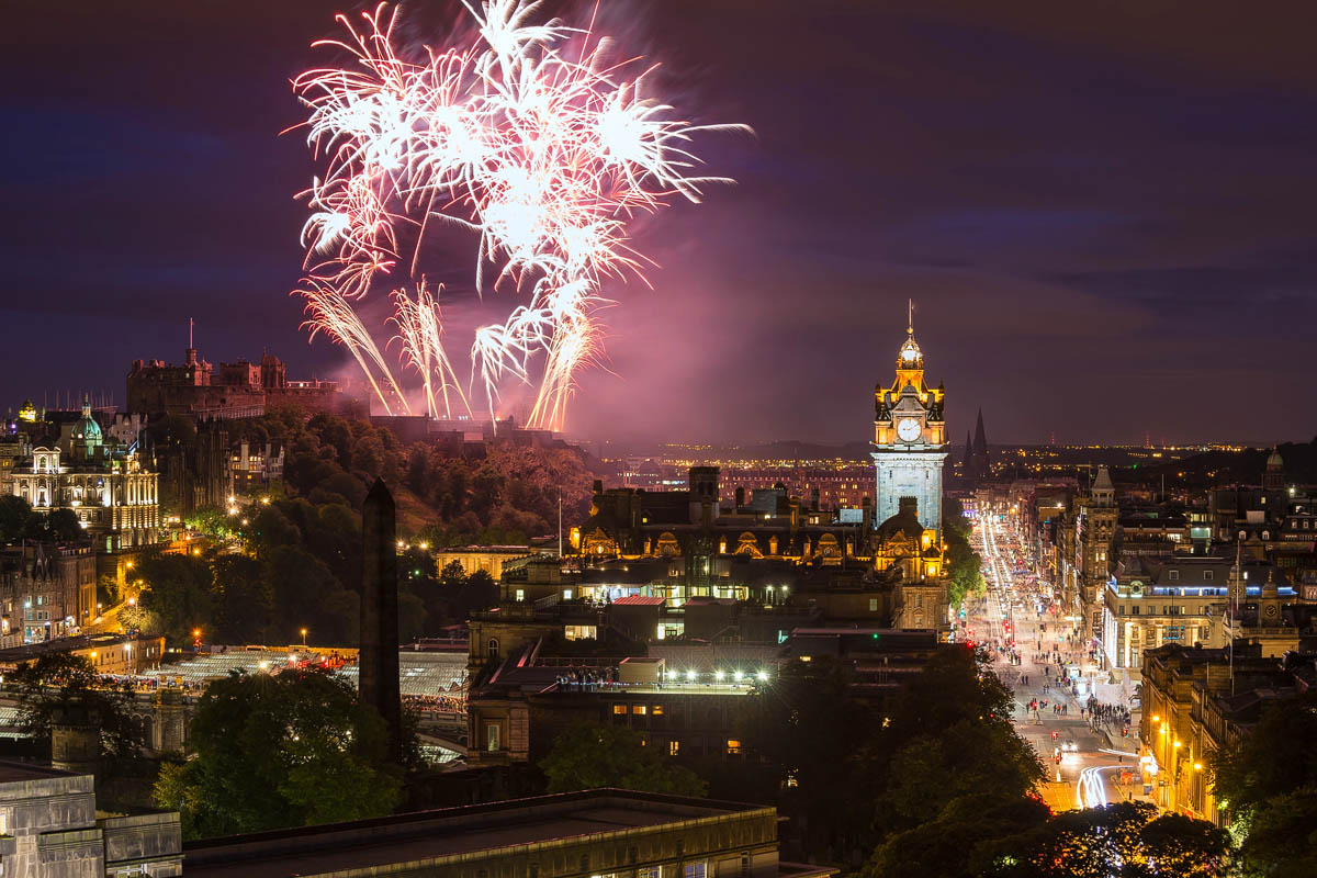 Fireworks exploding over Edinburgh Castle at night with the illuminated Balmoral Hotel clock tower and Princes Street below