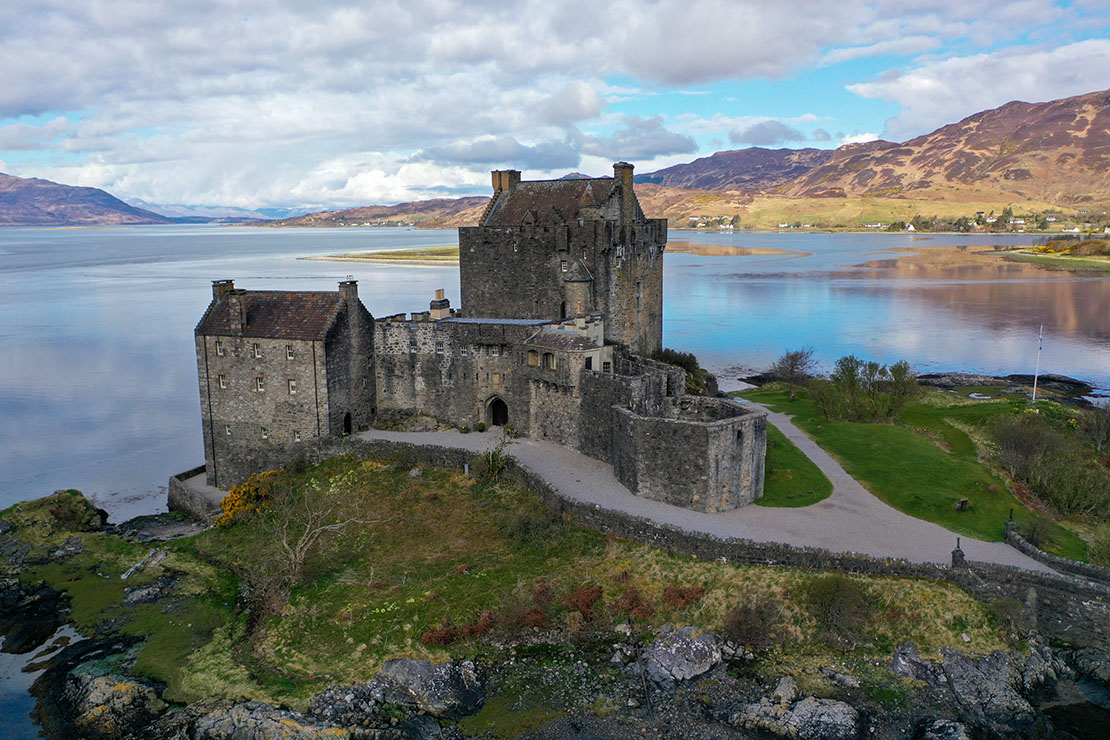 Aerial view of Eilean Donan Castle showing the keep, courtyard walls and main south entrance with loch and hills in the background on a calm spring morning