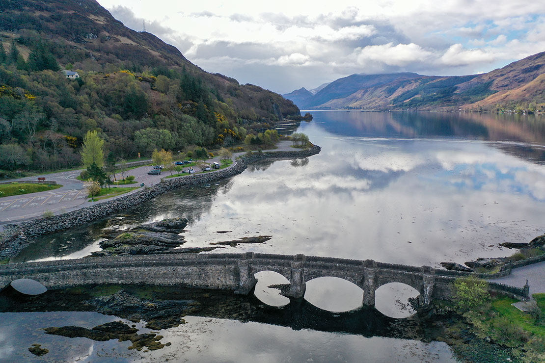 Aerial view of the Eilean Donan Castle footbridge leading to the visitor centre car park with the A87 road and Highland mountains in the background