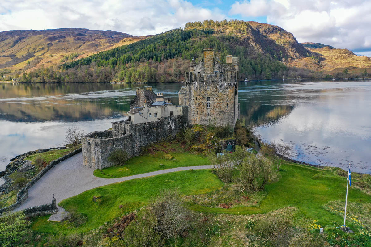 Close aerial view of Eilean Donan Castle with a Saltire flag flying on the lawn, daffodils in spring bloom, and the hornwork bastion visible