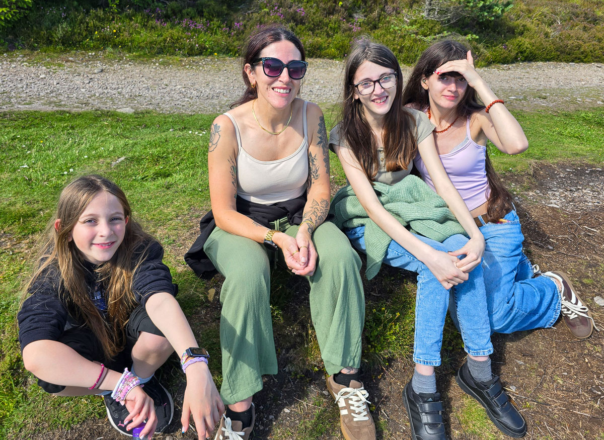 Janette and the three daughters sitting together on the grass at the monument, heather behind, all smiling