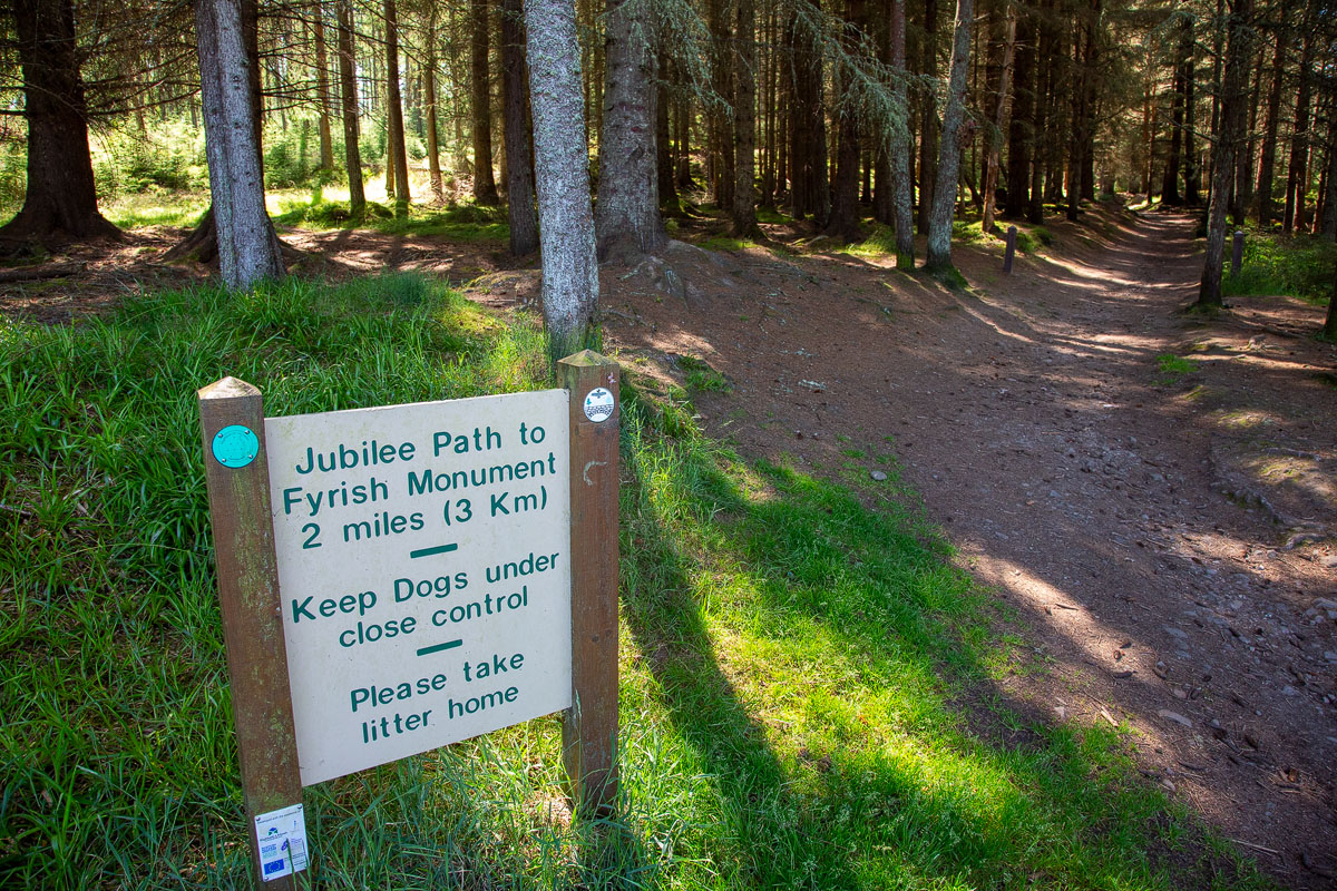 Green and cream Jubilee Path sign reading 2 miles 3km to Fyrish Monument, beside the wide forest track