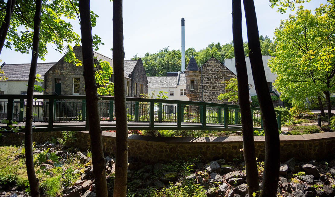 Glen Grant Distillery buildings in Rothes viewed through trees with the green footbridge crossing the burn in the foreground