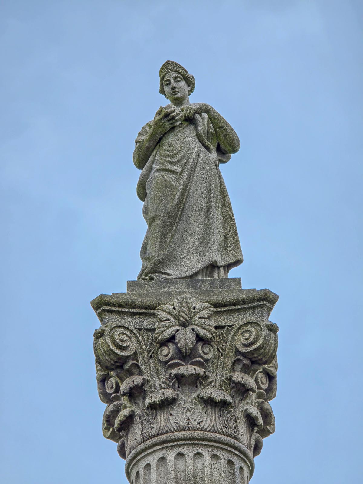 Close-up photograph of the granite statue of Hygeia in classical dress holding a cup, standing on an ornately carved Corinthian capital with scrollwork and acanthus leaves