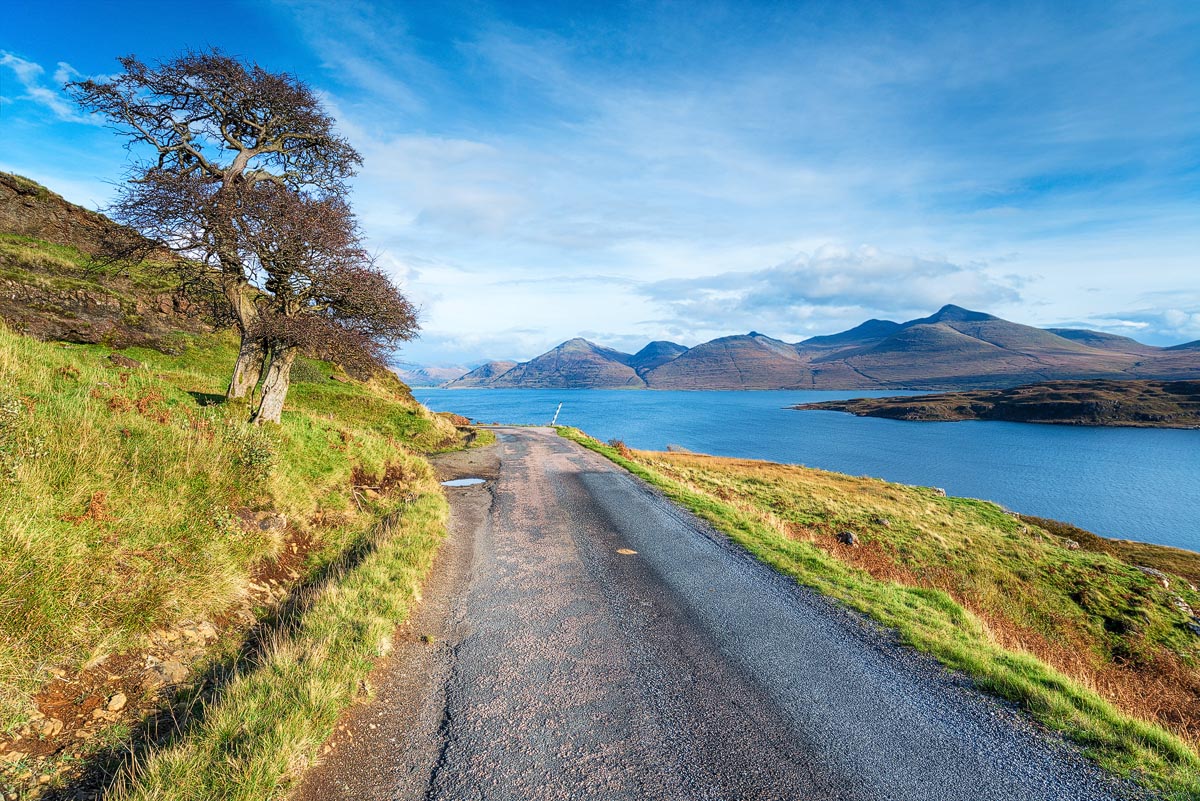 A single-track road on the Isle of Mull running alongside a sea loch with the Ben More mountain range in the distance