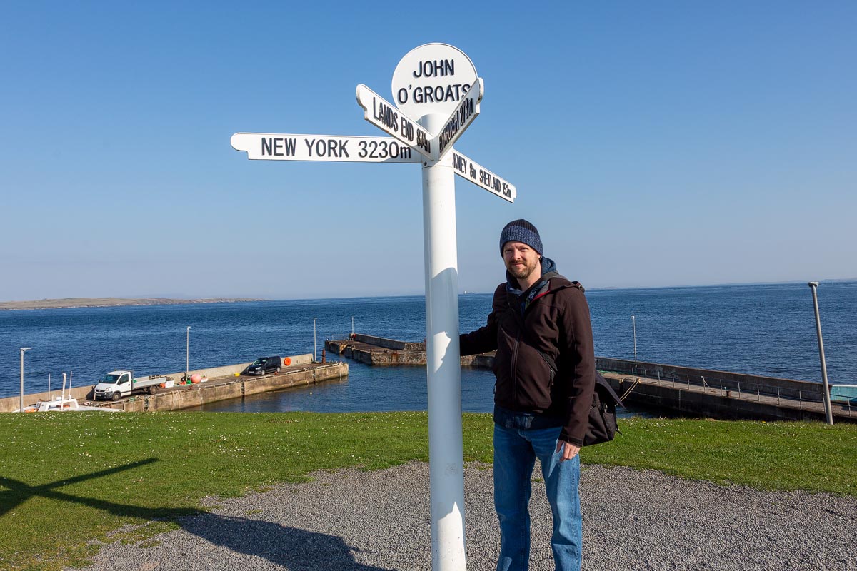 Chris standing at the John o Groats signpost with the harbour and Pentland Firth behind on a sunny day