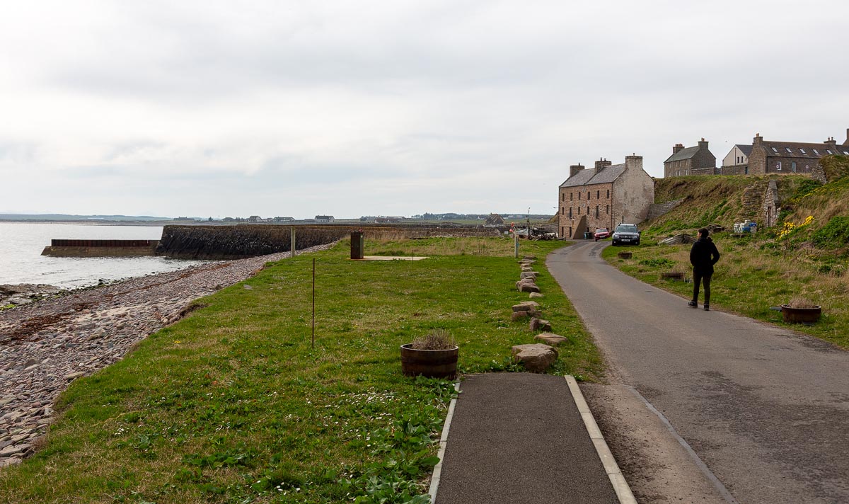 Walking down the road towards Keiss Harbour with the harbour wall and stone buildings visible