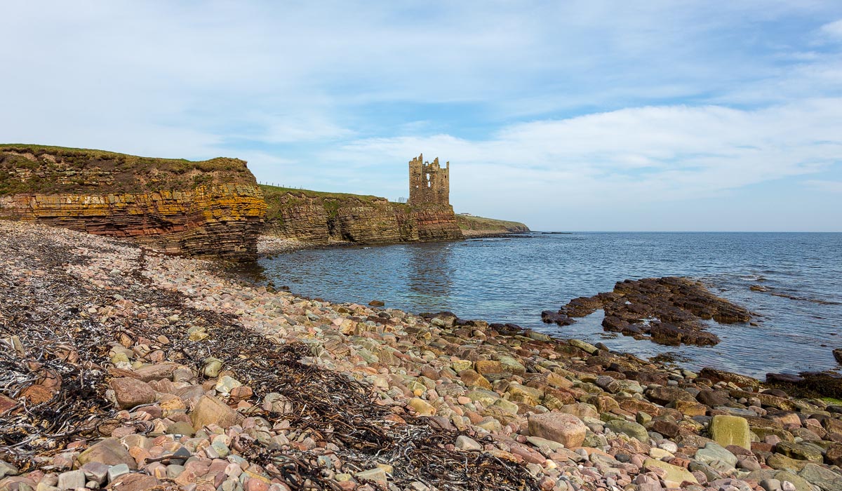 Wide view of Old Keiss Castle from the beach showing colourful stones, seaweed, and the castle perched on the cliffs