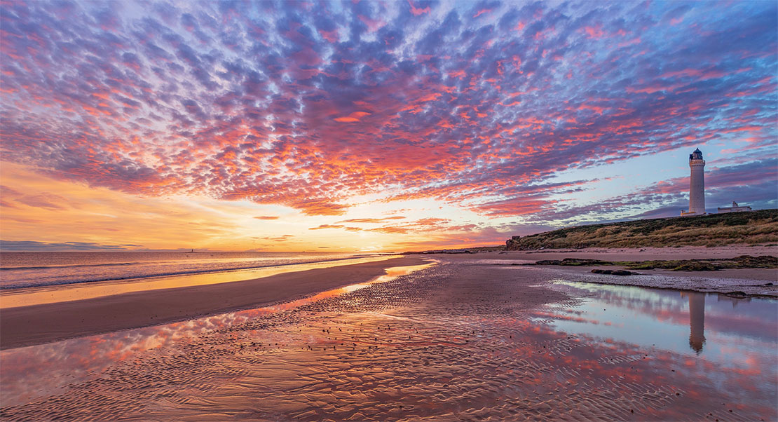 Lossiemouth West Beach at sunset with a dramatic mackerel sky reflected in wet sand and Covesea Lighthouse in the distance
