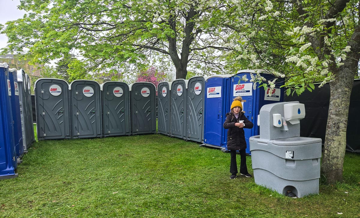 Row of grey and blue GAP portable toilets under trees with hand-wash station and Lauren in yellow bobble hat