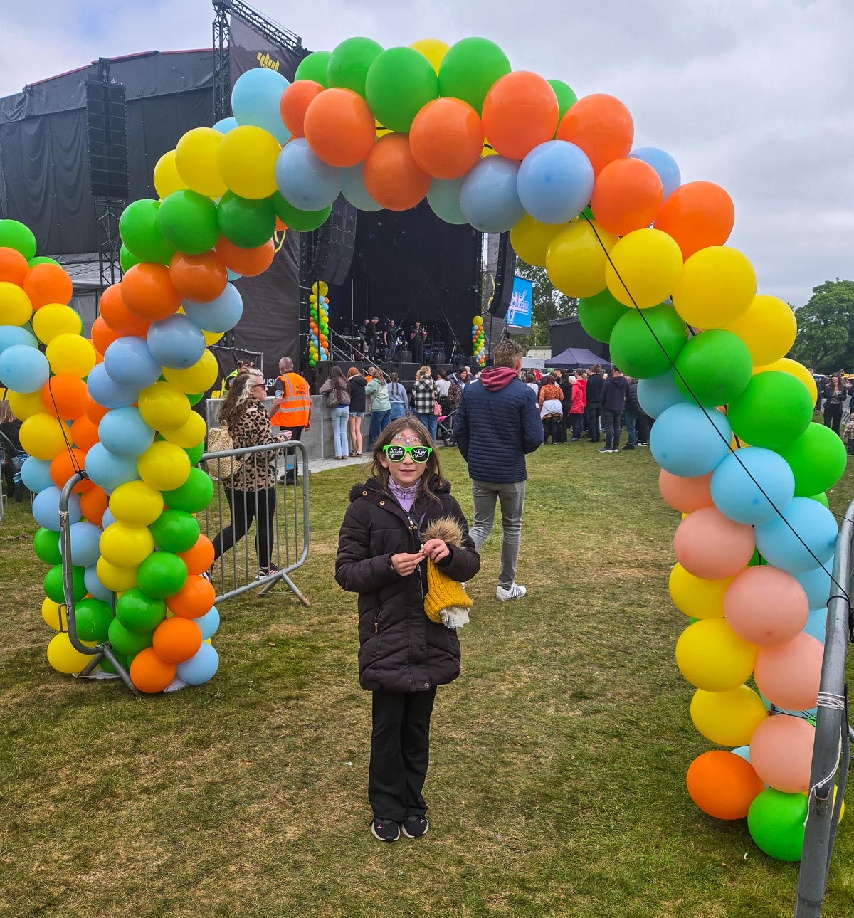 Young girl in green MacMoray sunglasses and face jewels standing under a colourful balloon archway in front of the main stage