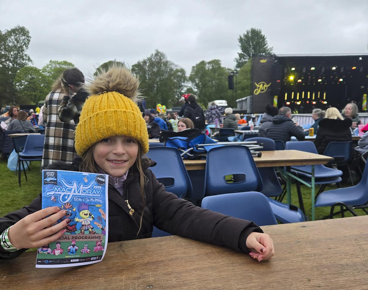 Young girl in yellow hat holding the 2026 MacMoray Festival official programme at a VIP table with the main stage in the background