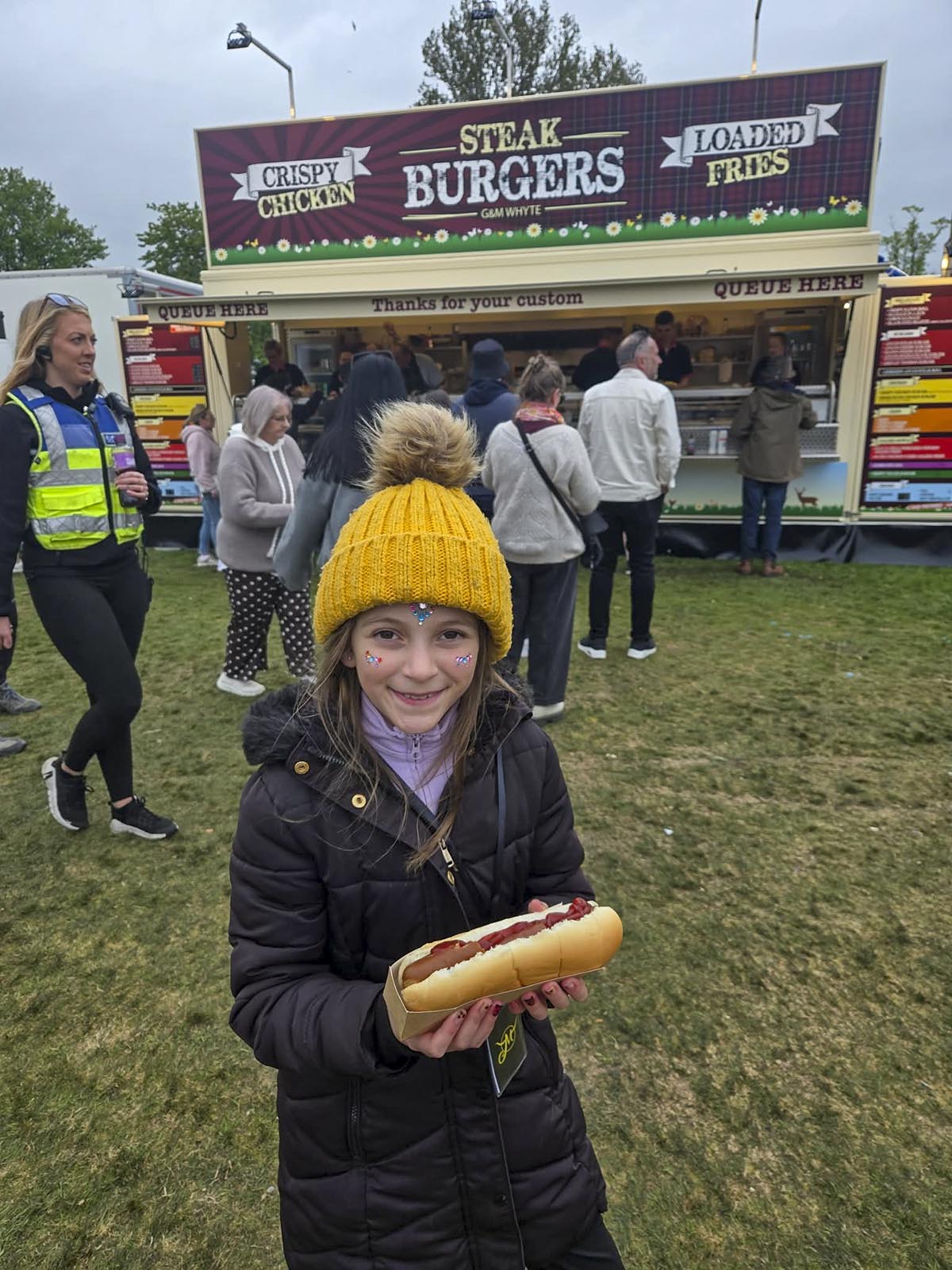 Young girl in yellow bobble hat holding a foot-long hotdog in front of the Steak Burgers food stand
