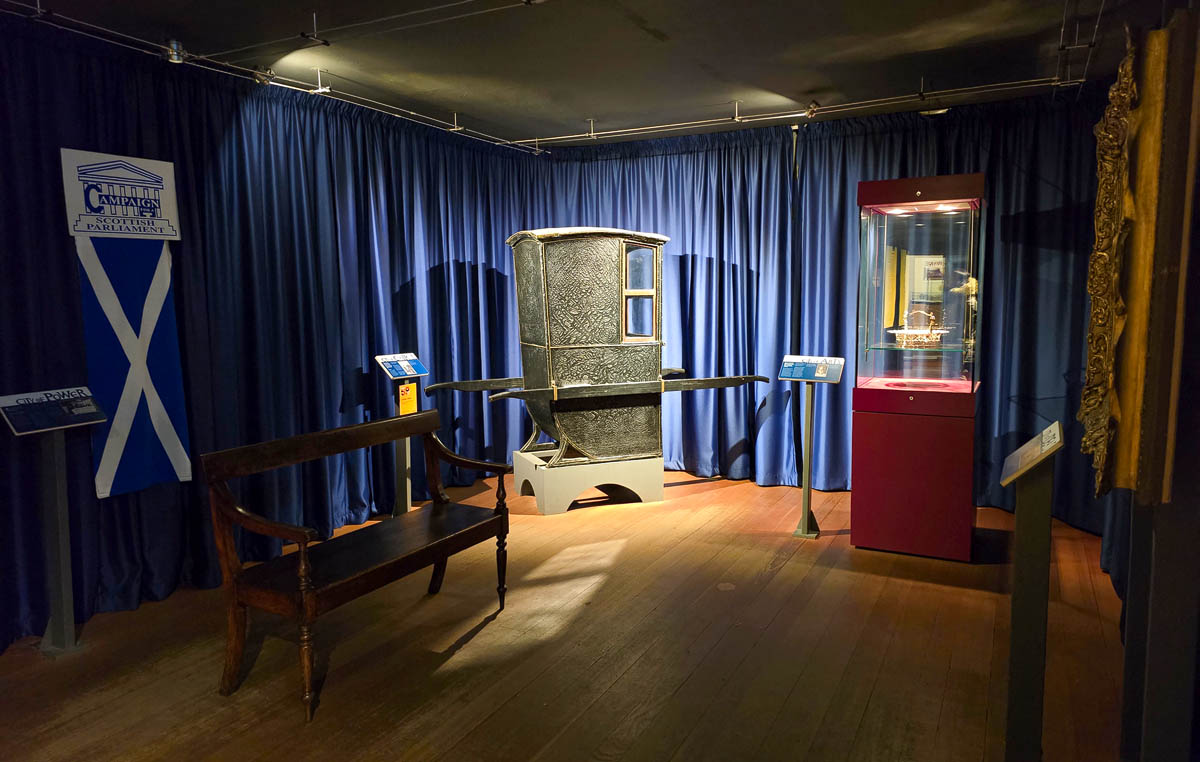 A sedan chair on display in a room with blue curtains, a Campaign for a Scottish Parliament sign and Saltire flag at the Museum of Edinburgh