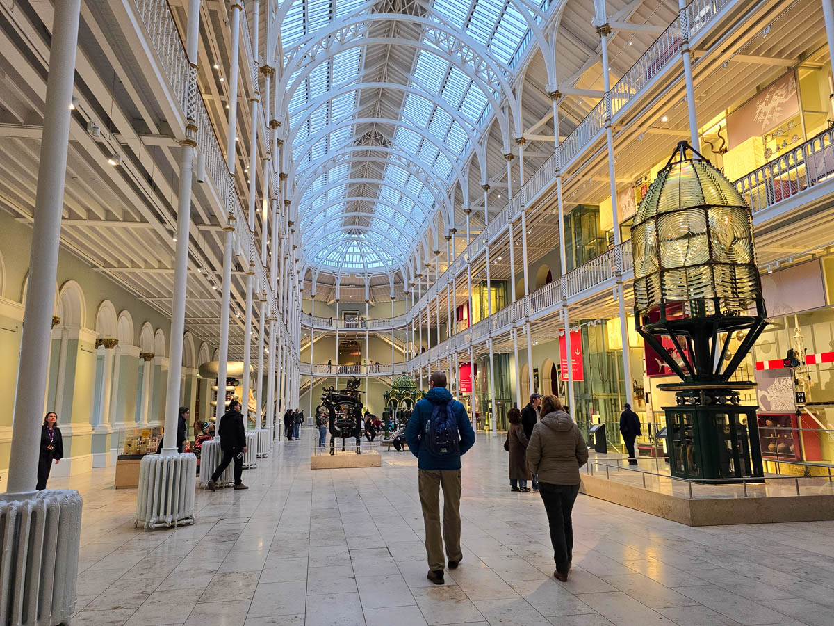 Visitors walking through the Grand Gallery at ground level with the Millennium Clock visible in the distance and a large lighthouse lens on the right