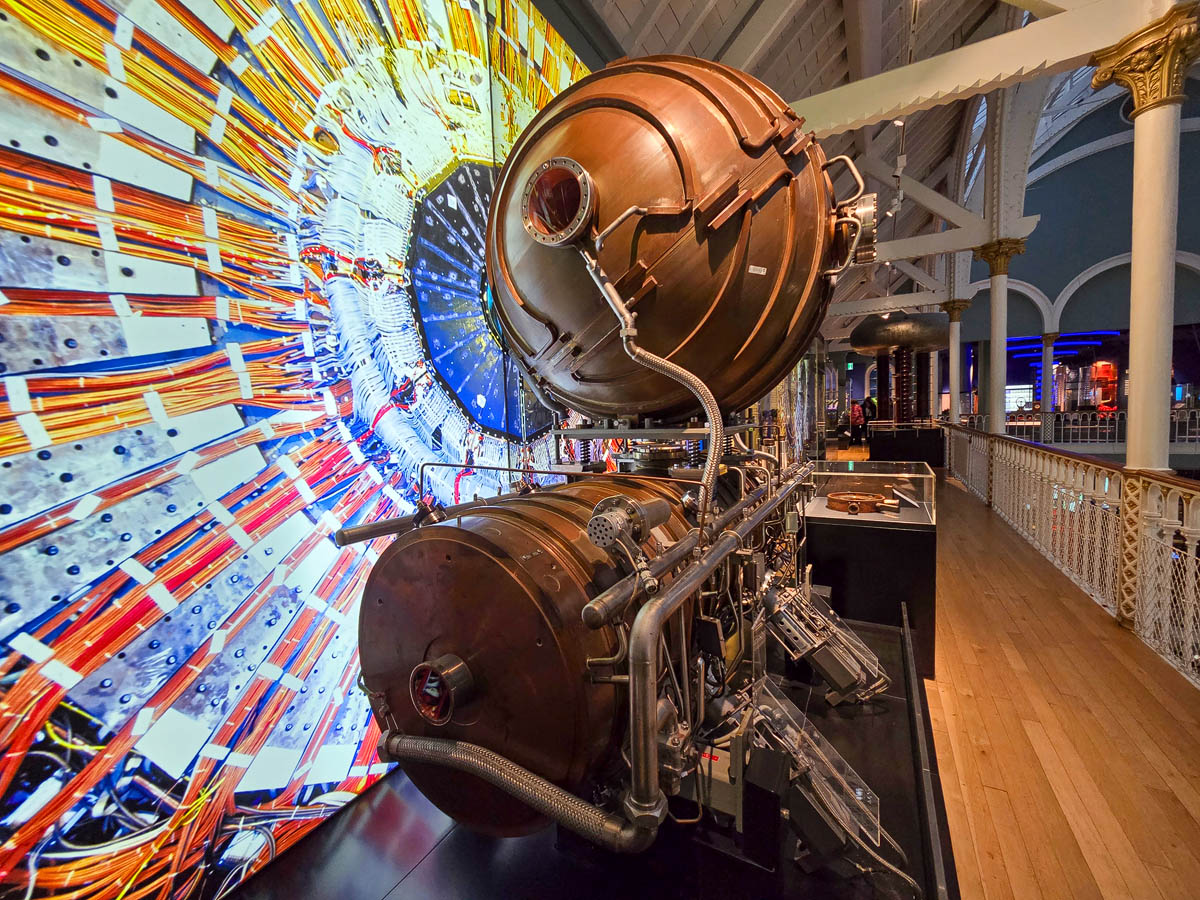 A large copper particle physics apparatus from CERN displayed against a colourful particle collision backdrop