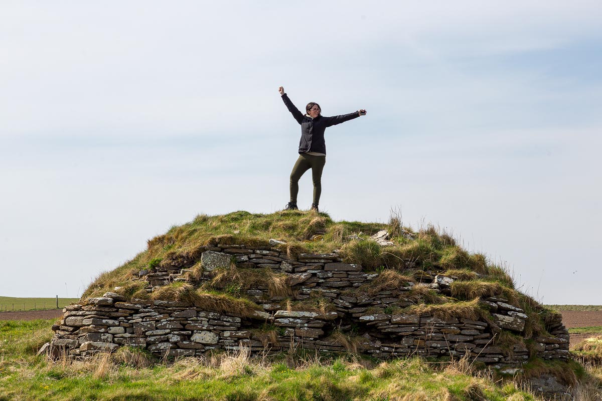 Janette standing triumphantly atop the grass-covered broch walls at Nybster with arms outstretched