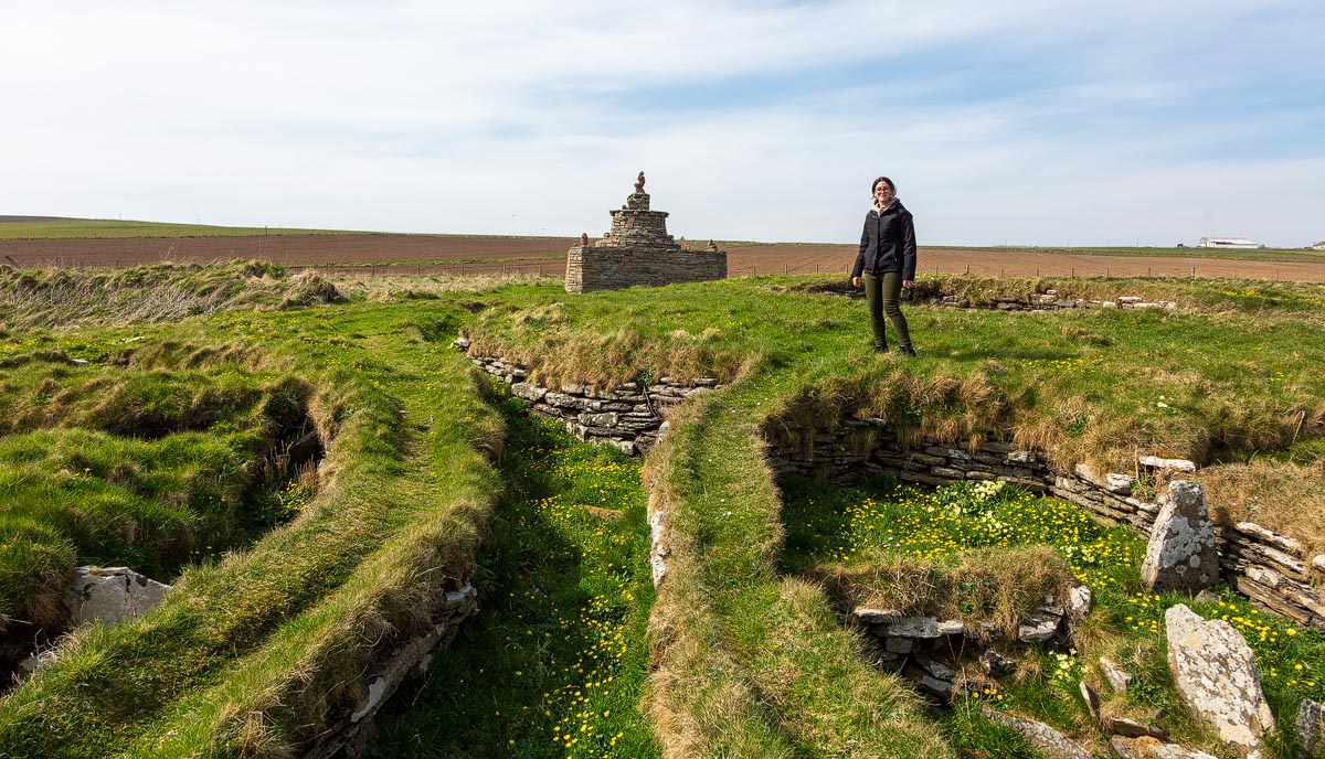 A visitor exploring the Nybster Broch settlement ruins with connecting passages and Mervyn’s Tower visible in the background