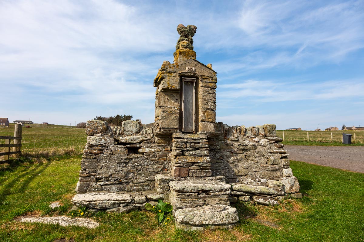 A carved stone memorial by John Nicolson near the Nybster Broch car park with a grotesque figure on top and date stone