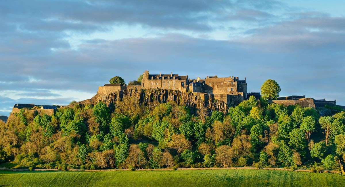 Stirling Castle bathed in golden evening light, perched on its volcanic crag above green woodland