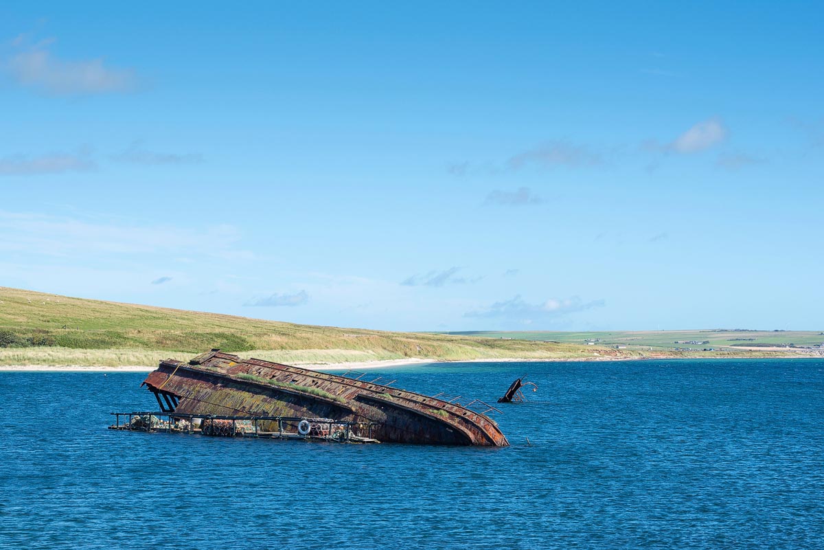 A rusting blockship wreck partially submerged in the calm blue waters near the Churchill Barriers