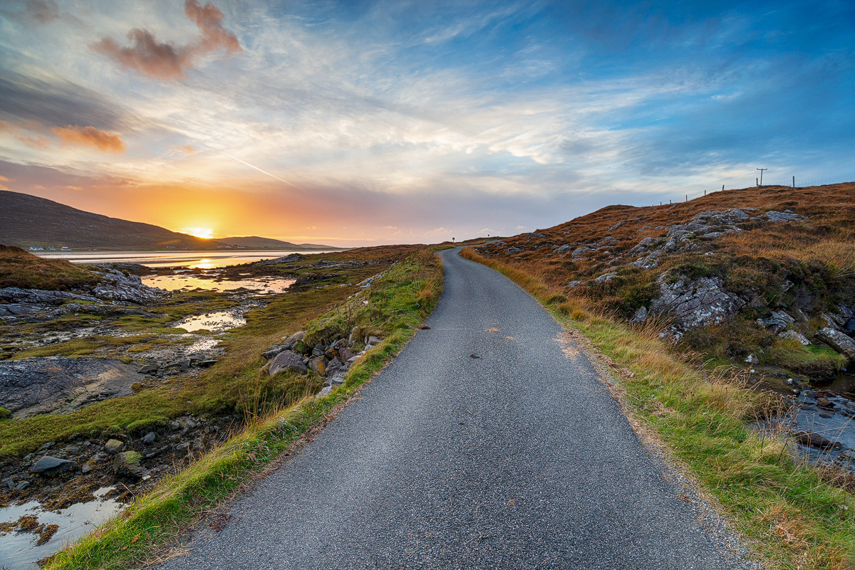 Single-track road winding along a rocky coastline towards the sea at sunset