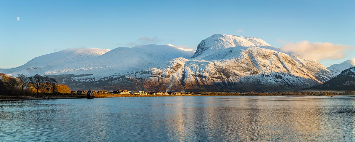 The snow-covered Ben Nevis range reflected in the calm waters of Loch Linnhe with a crescent moon overhead