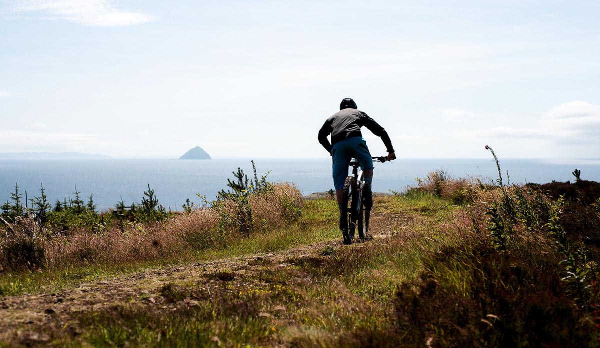 A mountain biker riding a coastal trail in Scotland with Ailsa Craig visible in the distance