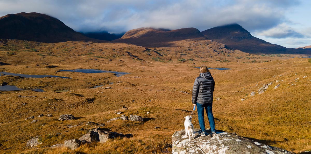 A walker with a small dog on a lead standing on a rock overlooking the Scottish Highlands