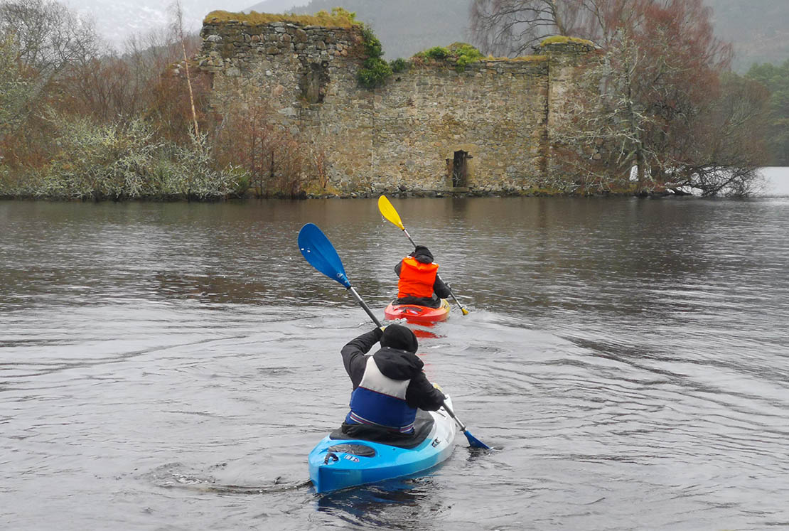 Two kayakers paddling on a Scottish loch towards a ruined castle on the shore