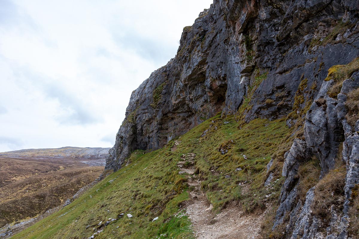 The steep approach path beneath the towering limestone cliff face of Creag nan Uamh with a cave entrance just visible